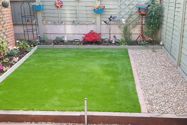 Small back garden with a rectangular artificial grass lawn edged in timber, a gravel strip with a block edge and a planted border against painted fence panels