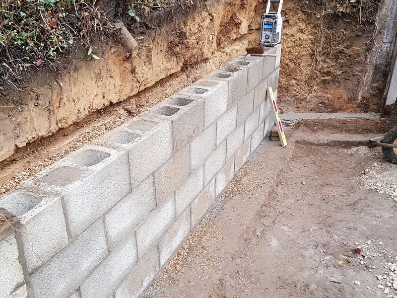 Concrete block retaining wall being built against a cut earth bank, with a spirit level on the top course