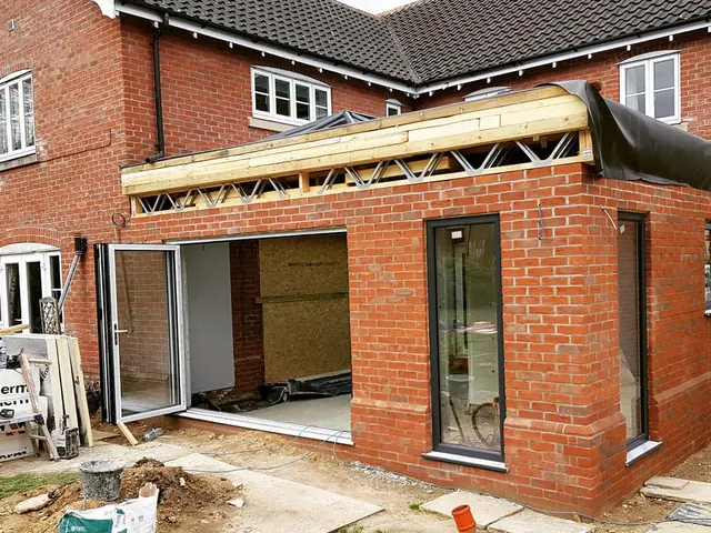 Single-storey red brick extension under construction at the back of a brick house, with flat roof joists in place and door and window frames fitted