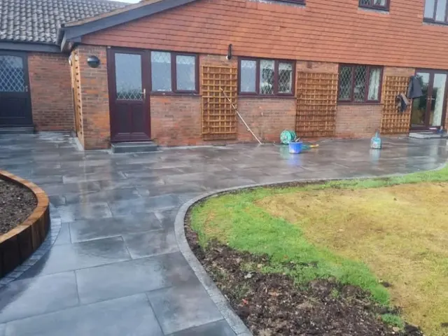 Dark grey paving sweeping in a curve around a lawn at the back of a brick house, with a timber sleeper raised bed on the left