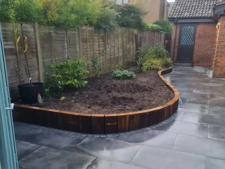 Curved raised planting bed built from upright timber sleepers, set into dark grey paving beside a fence