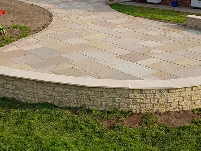Raised sandstone patio held by a low curved stone wall with a flat coping, seen from the lawn below