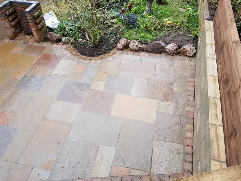 Indian sandstone patio in mixed buff, pink and grey slabs with a red block edge, a curved bed edged in rocks and a low block and sleeper retaining wall