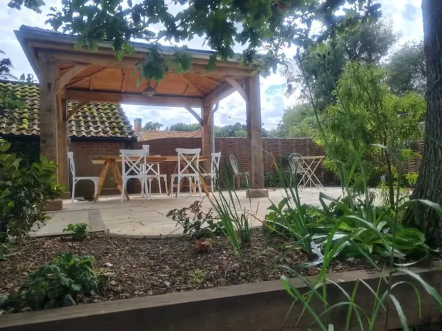 Oak-framed garden gazebo over a sandstone patio with white chairs, woven willow fencing behind and a planted bed with a timber edge in front