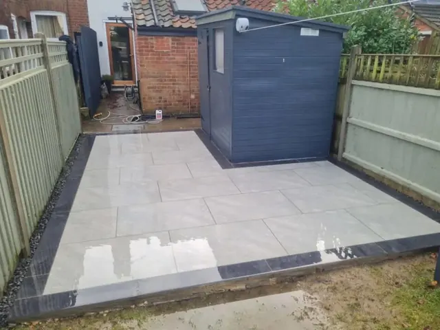 Pale grey porcelain patio with a dark border laid beside a grey garden shed, between two painted fences behind a terraced house