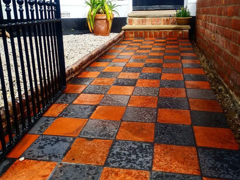 Red and black quarry tile path in a chequerboard pattern leading to a brick step and a dark front door, beside a brick wall and an iron gate