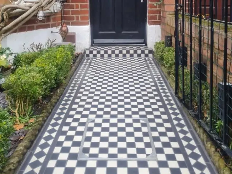 Black and white chequerboard tiled front path with a tiled border leading to the door of a red brick terraced house, with box hedging and iron railings